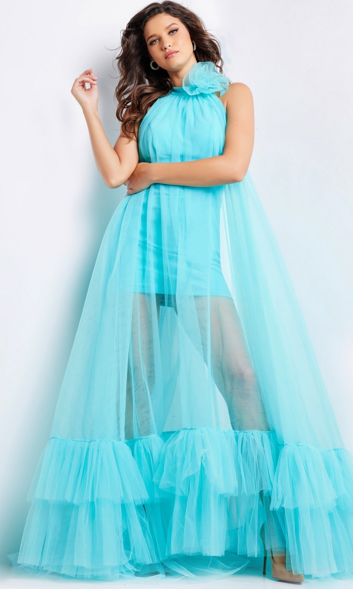 Woman wearing the Long Prom Dress 38720 by Jovani, a sleeveless turquoise tulle dress with sheer ruffled overlay and high neckline, posing against a plain white background.